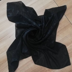 May include: A square black scarf made of a lightweight, semi-sheer fabric featuring a subtle, tonal jacquard pattern of abstract, flowing lines. The scarf is arranged in a central spiral fold, creating a layered, textured appearance across the surface.