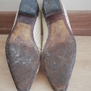 May include: A pair of off-white leather shoes, viewed from the soles. The soles are worn and show signs of use. The heels are dark gray and attached with small nails. The uppers have a woven pattern.