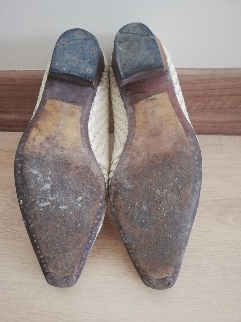 May include: A pair of off-white leather shoes, viewed from the soles. The soles are worn and show signs of use. The heels are dark gray and attached with small nails. The uppers have a woven pattern.