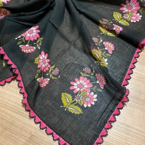 May include: A black scarf with a floral pattern featuring pink and white flowers and green leaves. The scarf has a pink crocheted trim around the edges, adding a decorative touch. The fabric appears lightweight and semi-transparent.