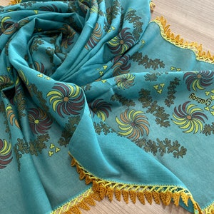 May include: A teal scarf with a floral pattern in brown, yellow, and olive green. The scarf has a yellow crocheted trim. The word "Guesoy" is printed on the scarf.
