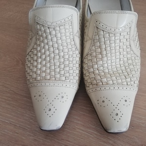 May include: A pair of off-white leather slip-on shoes. The upper features a woven design, with a smooth leather toe cap adorned with decorative perforations. The shoes have a pointed toe and a low heel.