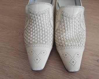 May include: A pair of off-white leather slip-on shoes. The upper features a woven design, with a smooth leather toe cap adorned with decorative perforations. The shoes have a pointed toe and a low heel.