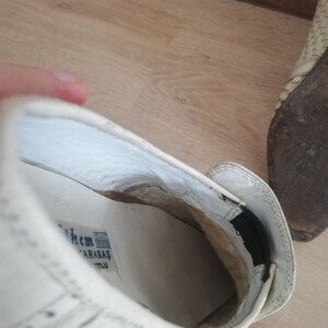 May include: Close-up of a white shoe with a woven pattern. The interior reveals a white insole with text and a black elastic band. The shoe is on a wooden surface.