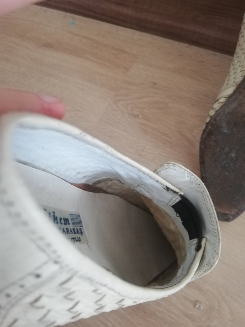 May include: Close-up of a white shoe with a woven pattern. The interior reveals a white insole with text and a black elastic band. The shoe is on a wooden surface.