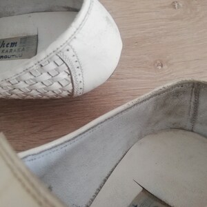 May include: Close-up of white leather shoes. The shoes have a woven design on the toe and a stitched detail along the side. The interior of the shoe is visible, with a label that reads "Ethem KARAKAŞ TURGUTLU".