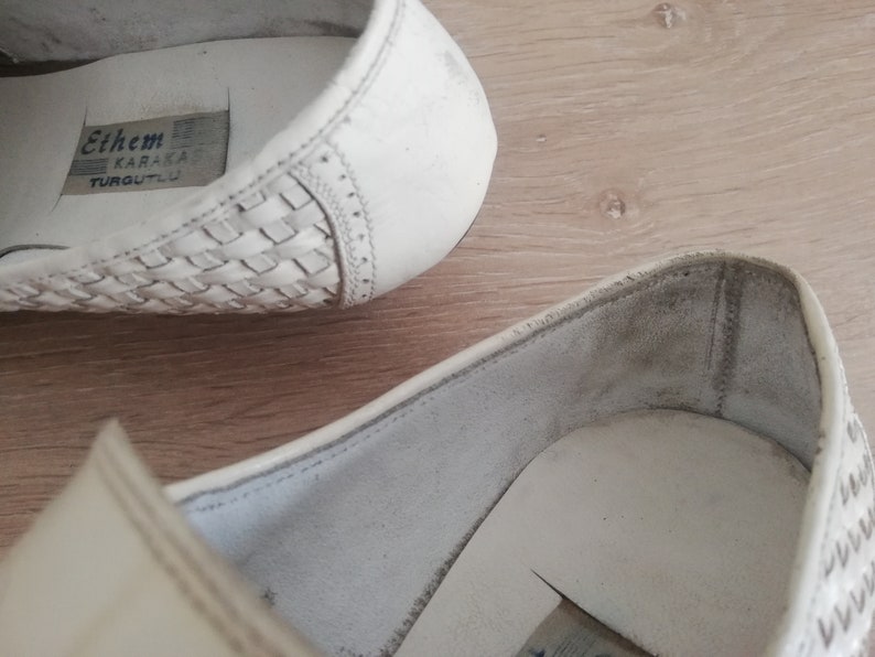 May include: Close-up of white leather shoes. The shoes have a woven design on the toe and a stitched detail along the side. The interior of the shoe is visible, with a label that reads "Ethem KARAKAŞ TURGUTLU".