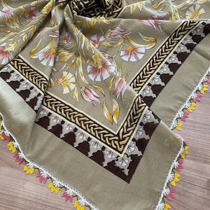May include: A beige square scarf with a floral print and a brown border. The scarf has a white crocheted trim with pink and yellow accents.