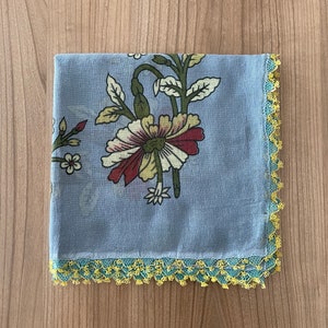 May include: A folded, square, light blue scarf with a floral pattern in red, white, and green. The edges are trimmed with a yellow and teal crocheted lace. The scarf is lying on a wooden surface.
