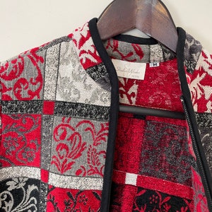 May include: A red, black, and white patterned jacket with a black zipper and a Studio Works Petite label. The jacket is hanging on a wooden hanger.