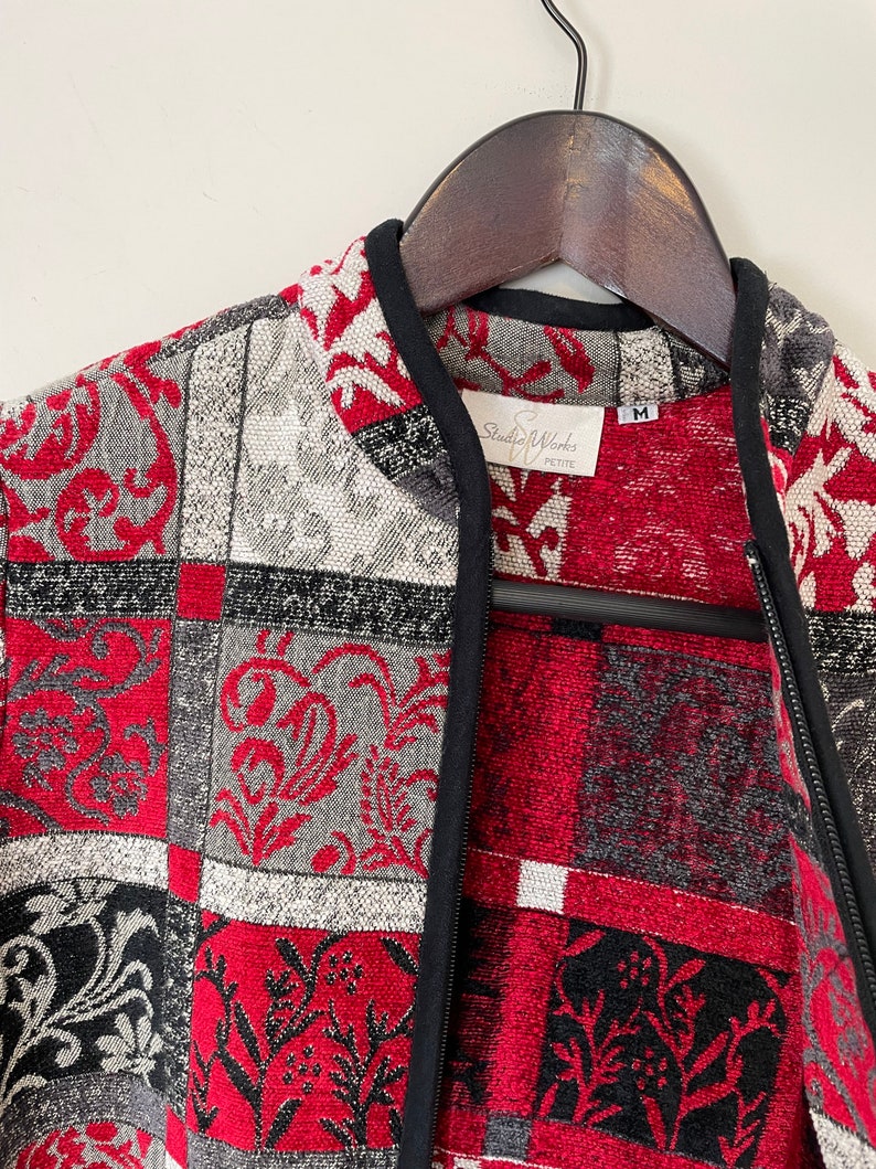 May include: A red, black, and white patterned jacket with a black zipper and a Studio Works Petite label. The jacket is hanging on a wooden hanger.