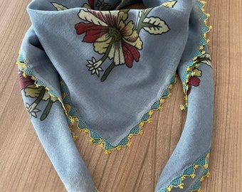 Pode incluir: Um lenço azul claro com um padrão floral em tons de vermelho, amarelo e verde. As bordas são adornadas com renda amarela e turquesa. Este lenço quadrado é um acessório elegante.