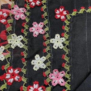 May include: Close-up of a black fabric adorned with delicate, handmade floral embroidery. The flowers feature red, pink, and white beaded centers, surrounded by green and red leaves. The edges are also embellished with floral patterns.
