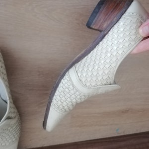 May include: Off-white woven leather shoe with a low heel and a pointed toe. The heel is a stacked wood design. The shoe has a woven pattern on the upper and a leather sole.