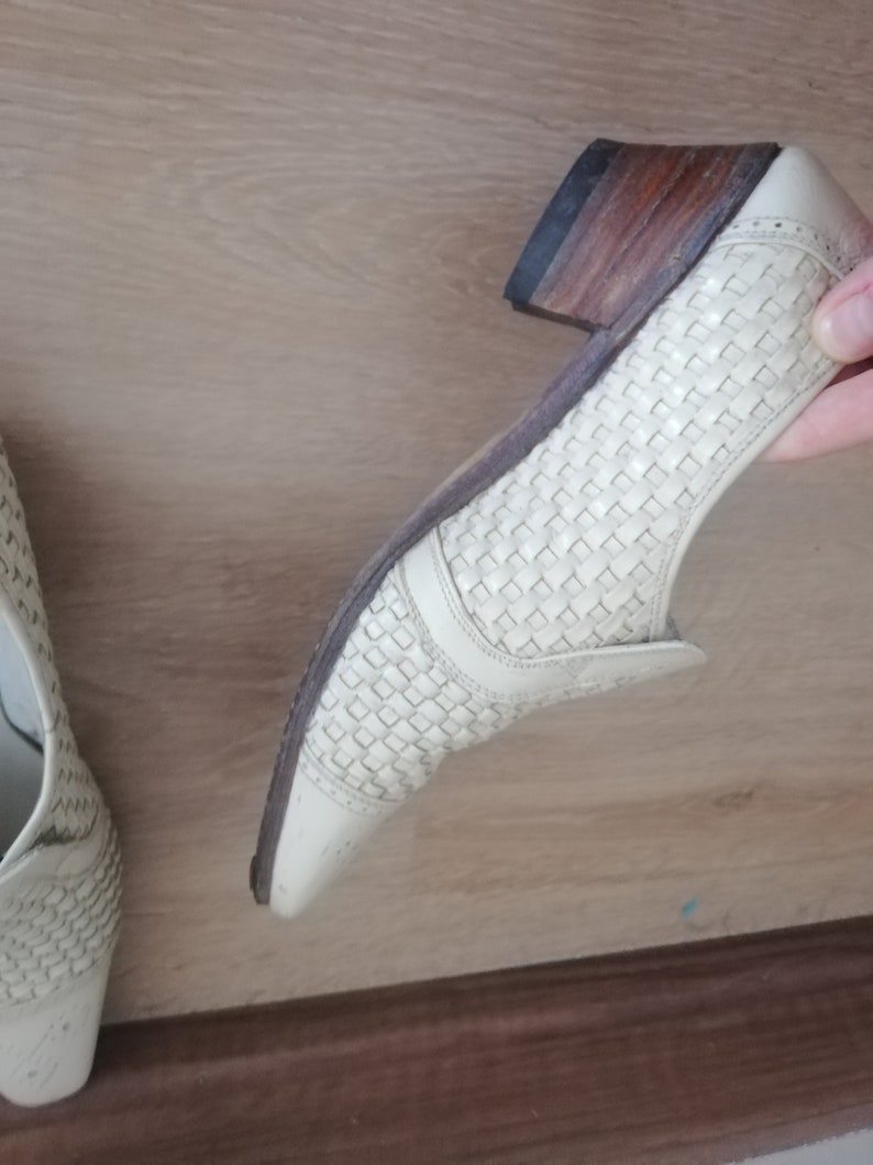 May include: Off-white woven leather shoe with a low heel and a pointed toe. The heel is a stacked wood design. The shoe has a woven pattern on the upper and a leather sole.