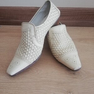 May include: A pair of off-white woven leather slip-on shoes. The shoes have a pointed toe with decorative perforations and a low heel. The woven pattern covers the top and sides of the shoes, creating a textured appearance.