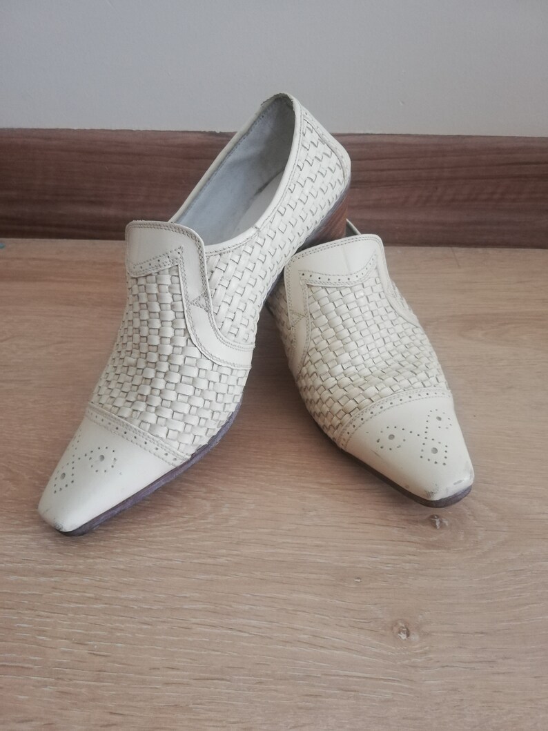 May include: A pair of off-white woven leather slip-on shoes. The shoes have a pointed toe with decorative perforations and a low heel. The woven pattern covers the top and sides of the shoes, creating a textured appearance.