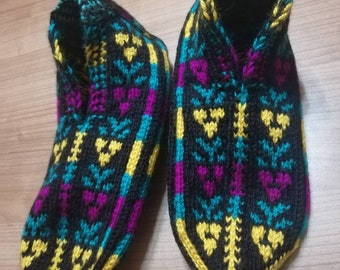 May include: A pair of knitted slippers with a black base, featuring a colorful pattern of yellow, teal, and purple geometric shapes. The slippers have a rounded toe and a cuff around the ankle. The soles are outlined in yellow.