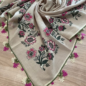 May include: A beige square scarf with a floral pattern in pink, white, and green. The scarf has a decorative border with green stitching and purple and white lace accents. The scarf is folded and draped, showcasing the design.