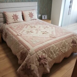 Pode incluir: Um edredom acolchoado rosa claro e duas fronhas a condizer com desenhos florais. A cama tem uma cabeceira cinza claro e está sobre um chão de madeira. Uma pequena telha decorativa está na mesa de cabeceira.