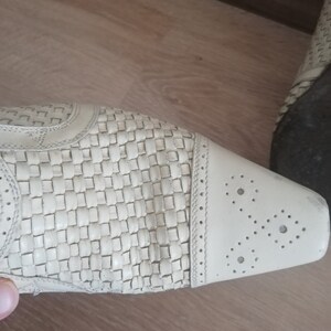 May include: Close-up of a cream-colored shoe with a woven pattern on the upper. The toe features decorative perforations. The shoe is on a wooden surface.