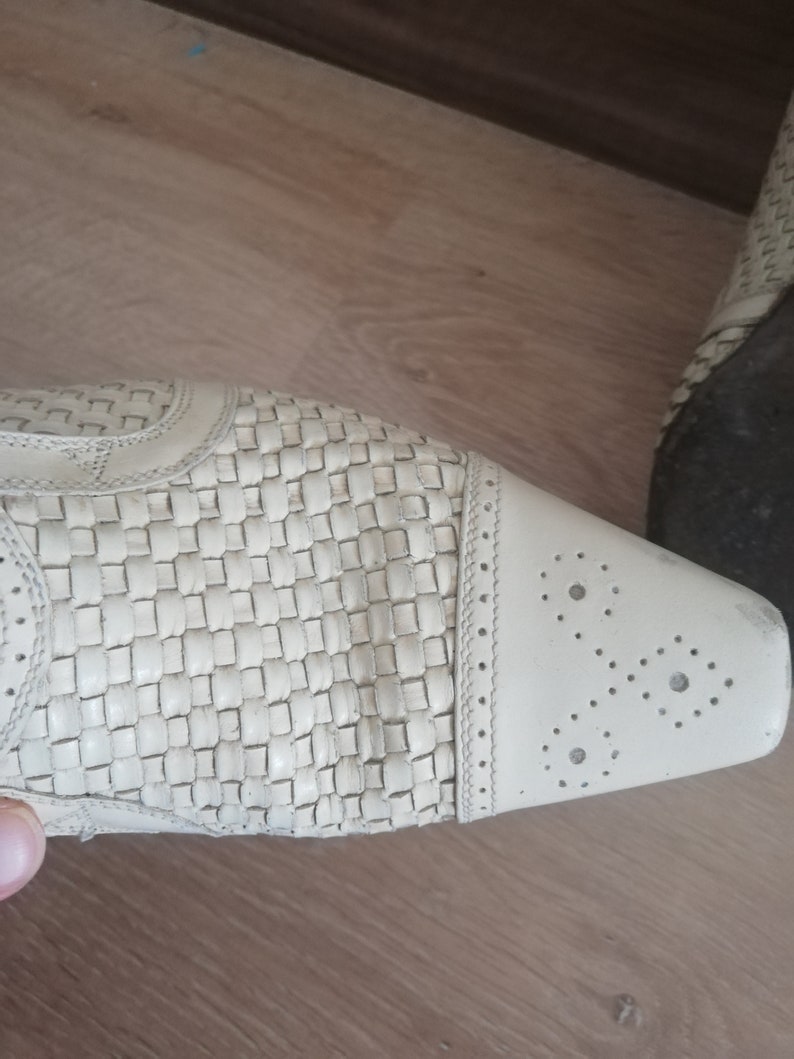 May include: Close-up of a cream-colored shoe with a woven pattern on the upper. The toe features decorative perforations. The shoe is on a wooden surface.