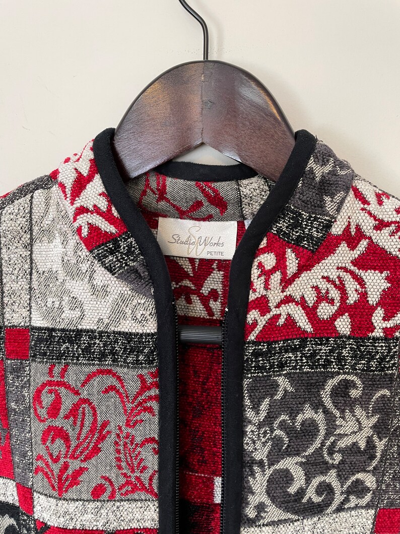 May include: A black zippered jacket with a patchwork design featuring red, gray, and white floral patterns. The label reads "Studio Works Petite."