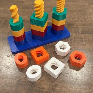 May include: A colorful wooden stacking toy with three vertical posts and various geometric shapes. The posts are topped with yellow screw-like shapes and sit on a blue base. Additional shapes include orange, white, and orange pieces.