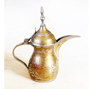 May include: A vintage brass and silver coffee pot with a long spout and a hinged lid. The pot is adorned with floral engravings and a decorative finial. The handle is curved, and the design suggests a Middle Eastern or North African origin. The pot is likely made of brass and silver.