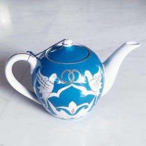 Peut inclure: Théière en céramique bleue et blanche avec un corps rond, une anse et un bec verseur blancs. La théière présente un bord argenté et un motif de deux colombes et d'anneaux entrelacés. Une ficelle bleue est attachée au couvercle.
