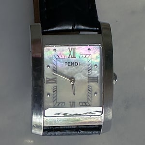 Può includere: Un orologio da polso Fendi rettangolare con quadrante in madreperla e cassa color argento. L'orologio presenta numeri romani e un cinturino in pelle nera. Il quadrante mostra il logo Fendi.