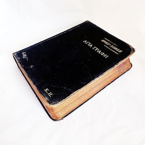 Op de afbeelding: Een zwart hardcover boek met gouden letters op de voorkant met de tekst HOLY BIBLE en AΓIA ΓPAΦH, en de initialen K.H. in goud in de onderste hoek. Het boek heeft verouderde, bruinachtige pagina's die zichtbaar zijn langs de zijkanten.