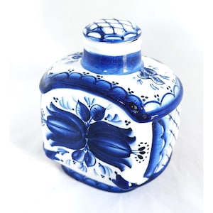 May include: A white ceramic container with a blue floral pattern and a matching lid. The container has a rounded shape with a cylindrical neck and a lid with a crosshatch design. The floral design features blue flowers and leaves.