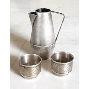 Puede incluir: Una jarra de metal plateado con asa y boquilla, acompañada de dos pequeñas tazas a juego. La jarra y las tazas tienen una banda decorativa alrededor de la base. El juego es probablemente para servir bebidas.