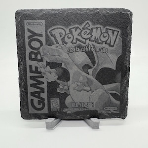 May include: A black slate coaster featuring the Pokémon Red Version Game Boy game design. The coaster has the Game Boy logo on the left and the Pokémon logo with a Charizard illustration in the center. The text "Gotta catch 'em all!" is also visible.