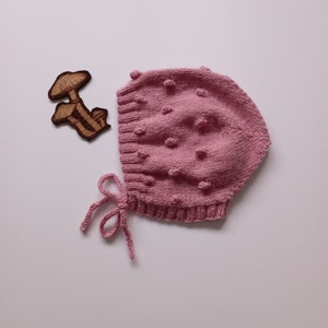 May include: A pink knitted baby bonnet with a textured bobble pattern and a tied ribbon.