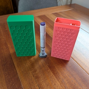 Op de afbeelding: Een groene rechthoekige koffer met een honingraatpatroon en een koraalkleurige koffer met een hartpatroon flankeren een medische injector. De items staan op een houten oppervlak.