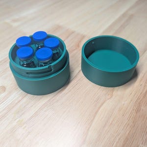 Può includere: Un contenitore circolare verde acqua con coperchio, contenente cinque piccole fiale di vetro con tappi blu. Il contenitore è aperto, rivelando le fiale all'interno. Il coperchio è posto di lato. Su una superficie di legno.