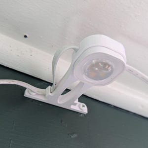 Puede incluir: Un foco LED blanco montado en una pared. El accesorio tiene una luz pequeña y redonda y se fija con un soporte y tornillos. El cable eléctrico es blanco.
