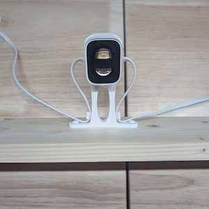 May include: A white security camera mounted on a white plastic stand with a rectangular lens housing. The device features a black interior frame around the lens and includes a white power cable extending from the base to the sides of the unit.