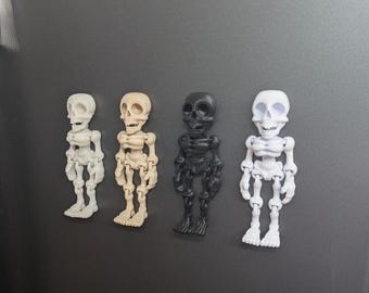 May include: Four small skeleton figurines in different colors: off-white, tan, black, and white. Each figurine is a detailed representation of a human skeleton, with articulated limbs and a skull. The figurines are arranged in a row.
