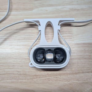 May include: A white plastic lighting fixture featuring a central vertical support with an oval cutout and a horizontal base. The light unit contains three distinct circular lenses set within a dark gray housing, with white power cords extending outward.