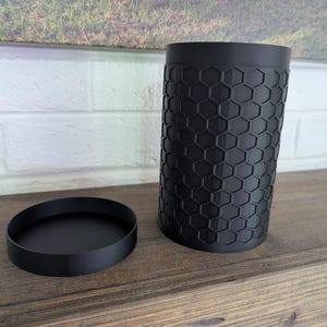 May include: A black cylindrical container with a matching round lid. The container has a textured surface with a honeycomb pattern. The objects are displayed on a wooden surface.