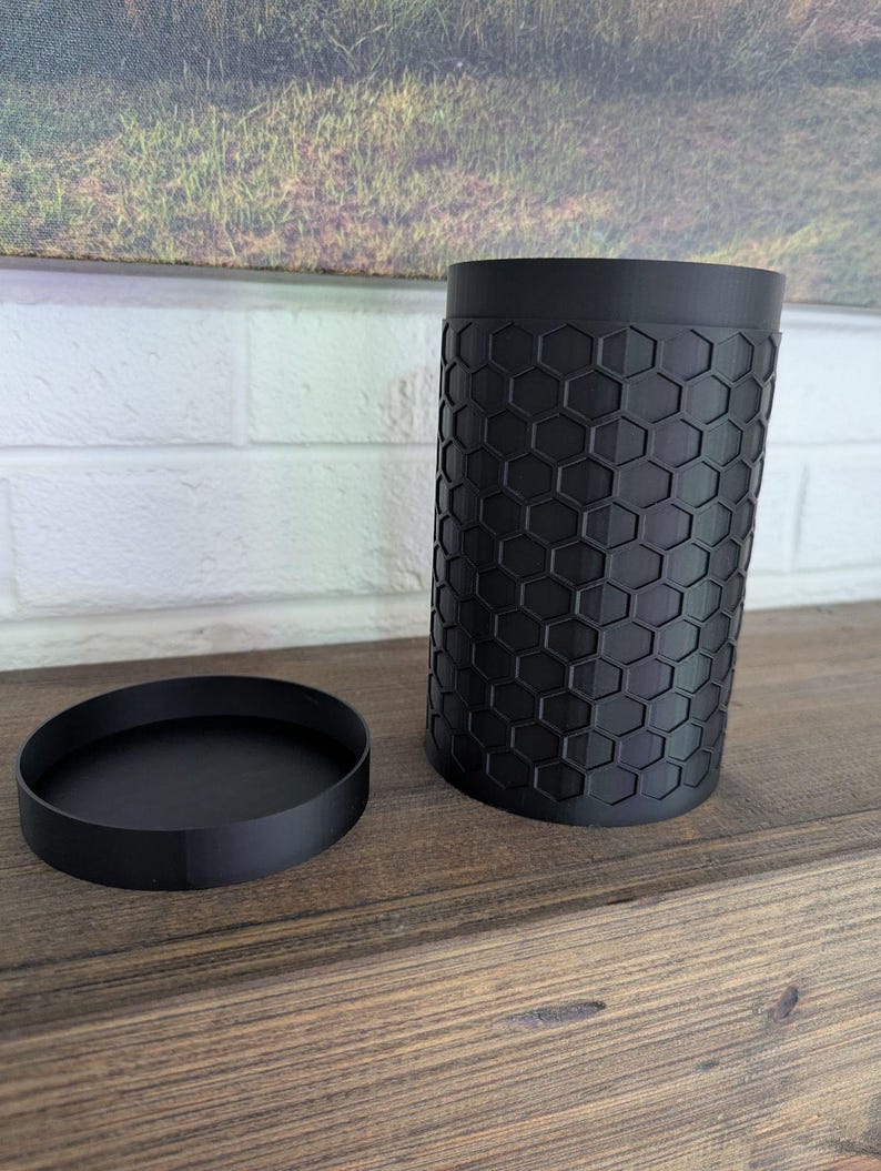 May include: A black cylindrical container with a matching round lid. The container has a textured surface with a honeycomb pattern. The objects are displayed on a wooden surface.