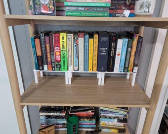 May include: A wooden bookshelf with multiple shelves, displaying a collection of books. The books are arranged on the shelves, with some supported by white book stands. The books have various colored spines and titles, creating a visually appealing arrangement.