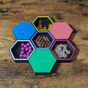 May include: A colorful hexagonal game organizer with six compartments, each holding different game pieces. The compartments are blue, yellow, pink, green, purple, and black, and contain various game tokens.