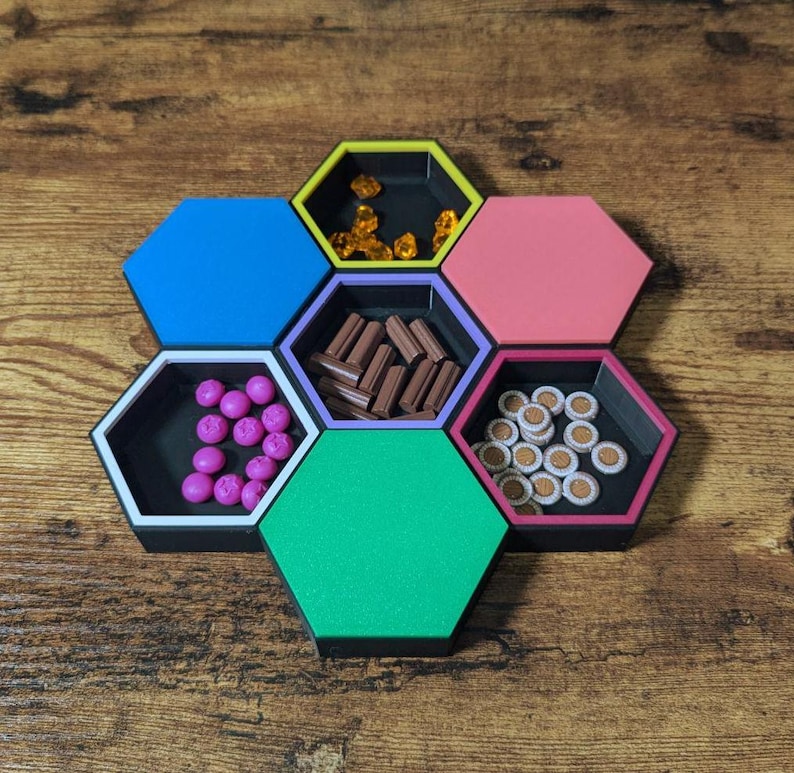 May include: A colorful hexagonal game organizer with six compartments, each holding different game pieces. The compartments are blue, yellow, pink, green, purple, and black, and contain various game tokens.
