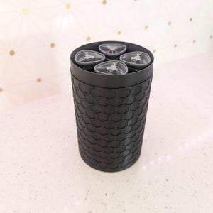May include: A black cylindrical container with a honeycomb pattern on the exterior. The top of the container holds four clear, gray-toned objects. The container is on a white speckled surface.