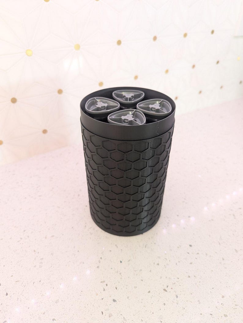 May include: A black cylindrical container with a honeycomb pattern on the exterior. The top of the container holds four clear, gray-toned objects. The container is on a white speckled surface.