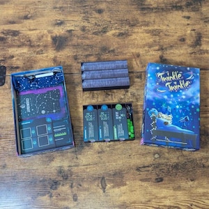 May include: A board game set on a wooden table. The game box is titled "Twinkle Twinkle" with a blue and white design. Other game components include a box with cards and a box with game pieces.